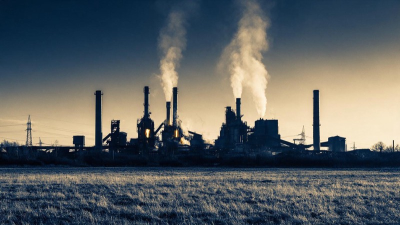 A steelworks at dusk with glowing furnace chimneys and pale plumes of steam rising into a still sky, shot across a flat field of winter grass