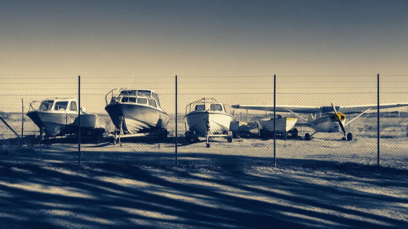 Surplus boats and a small propeller aeroplane parked side by side in a fenced yard on a dry plain, long shadows and wire fencing in the foreground