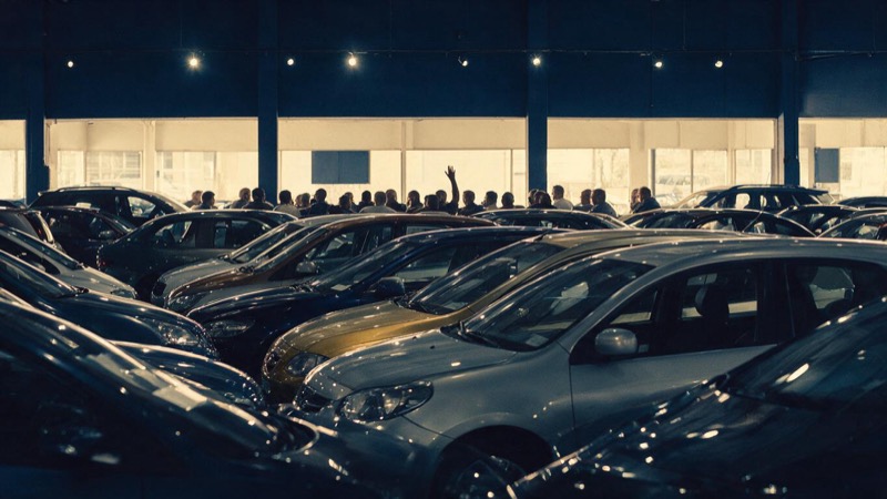 Wide shot of a salesroom hall with dozens of used cars in tidy rows under spotlights, a single raised hand silhouetted among seated buyers in the distance