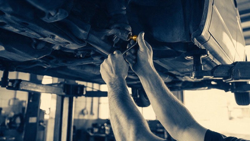 A van raised on a hydraulic lift in a service garage, a mechanic's arms and hands tightening a bolt beneath it, bright workshop light, close-up