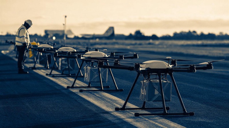 A row of small delivery drones on a launch field at dawn, rotors still and parcels slung beneath, a ground crew in hi-vis standing nearby