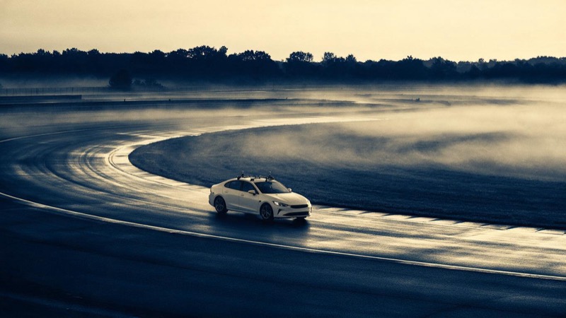 A white test sedan with roof-mounted sensors circles a wet proving-ground track at dawn, mist lying low across the infield, seen from a distance at the edge of the circuit