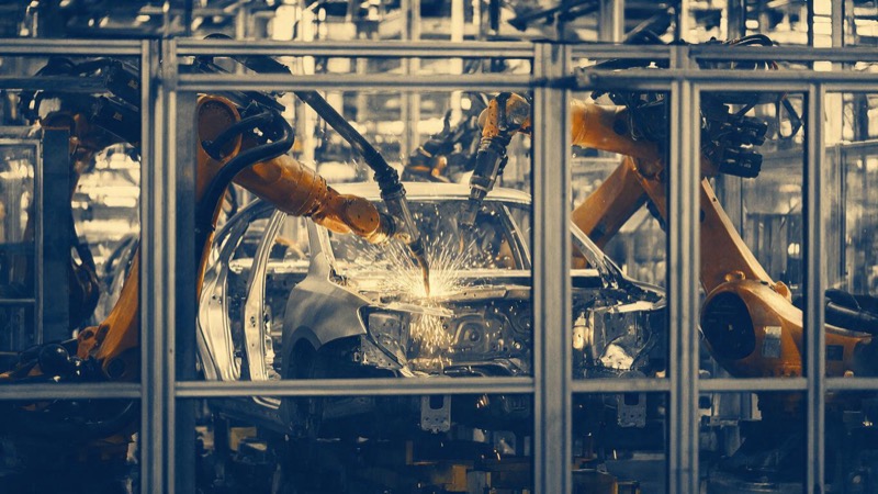 Orange industrial robot arms welding a car body shell in a Michigan plant, shot from outside a safety fence with a shower of sparks, no people in view