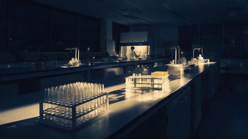 Wide view of a quiet laboratory at night with long benches, racks of test tubes catching the light and a single technician working far off at a safety cabinet