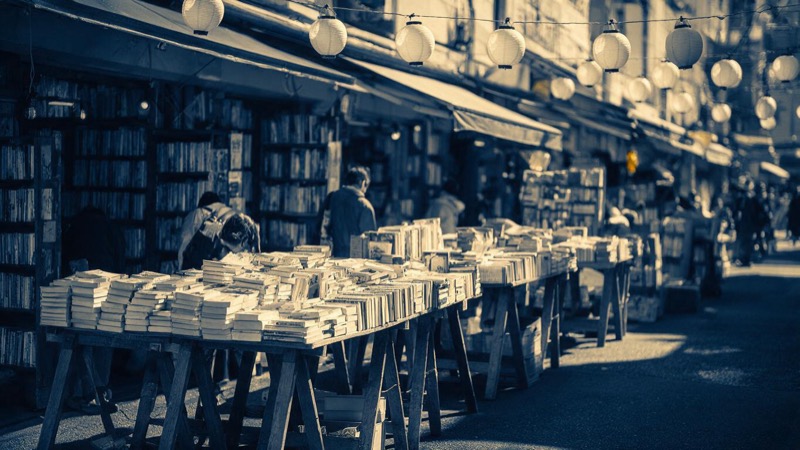 A street of second-hand bookshops in Tokyo at midday, trestle tables heaped with books outside every shop, small figures browsing, plain paper lanterns hanging overhead