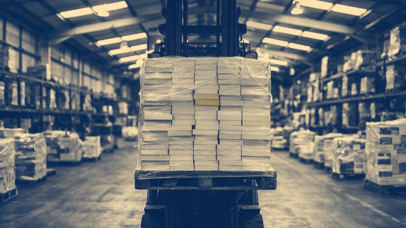 A pallet of shrink-wrapped identical paperbacks lifted by a forklift in a publisher's warehouse in the English Midlands, a long aisle running away beneath high strip lights