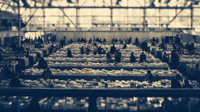 Wide view from a mezzanine of a vast book-fair hall in Frankfurt, small figures meeting at long rows of tables stacked with books, soft daylight pouring through a glass roof
