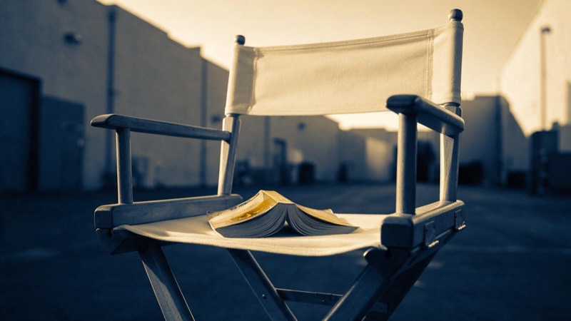 A dog-eared paperback lying open on the folded canvas of an empty director's chair on a quiet Los Angeles film backlot at golden hour, sound-stage walls beyond