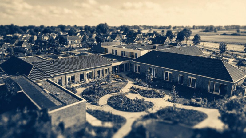 Aerial view of a low brick care campus of linked courtyards and garden paths on the edge of a Dutch town in soft afternoon light