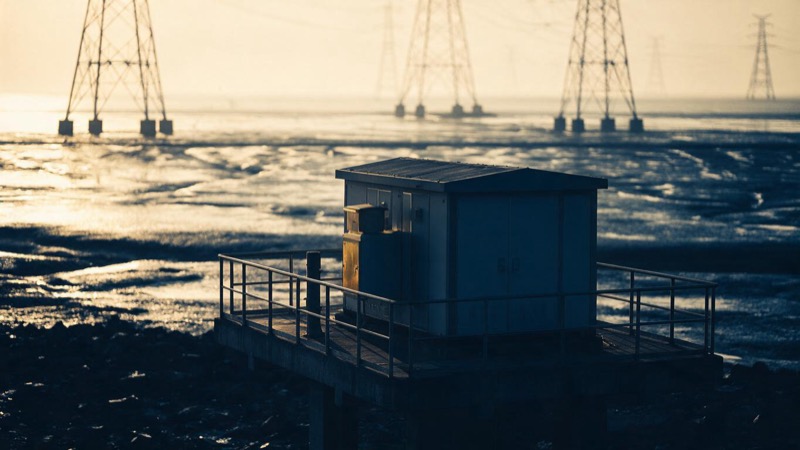 A low-lying coastal substation standing on a raised concrete platform above wet tidal flats at sunrise, pylons receding into haze