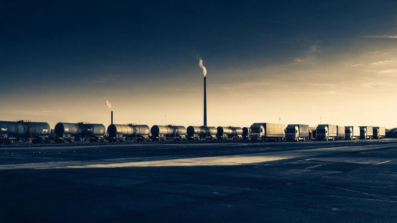 Wide dusk view of a freight yard where a train of tank wagons, a row of lorries and a distant chimney with a thin plume share one flat industrial horizon