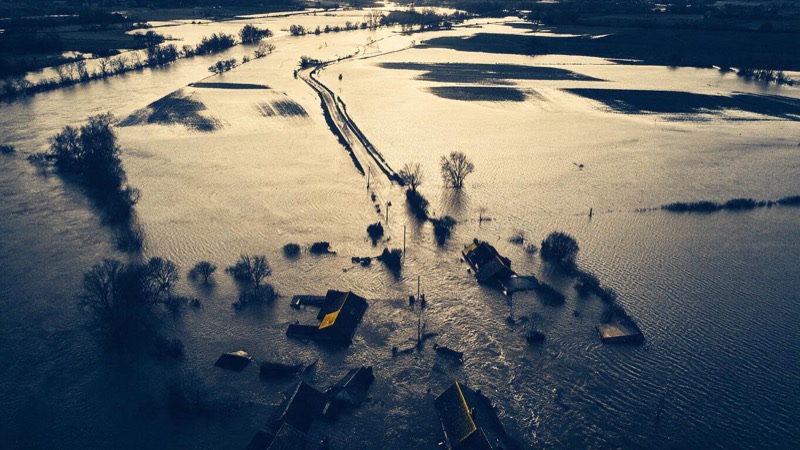 Aerial view of a flooded river valley after a storm, brown water covering fields and a village road with rooftops showing above the surface in pale morning light