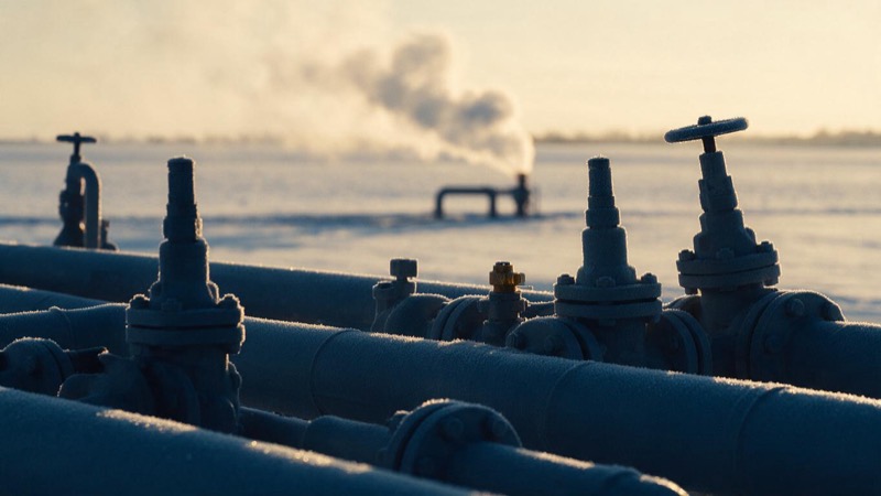 Frosted pipework and valves at a gas storage site in northern Germany at pale winter dawn, steam rising from a vent across a flat white field