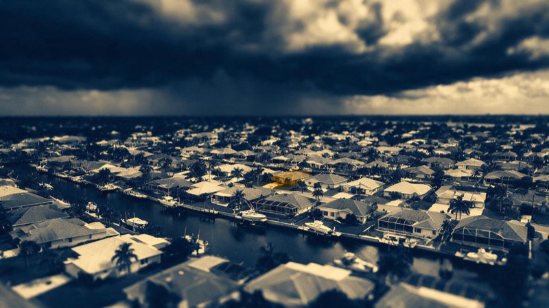 Aerial view of a Florida canal neighbourhood of similar rooftops and moored boats under a bruised storm sky, the water unnaturally still
