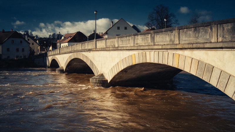 A swollen brown river running fast beneath a stone bridge in a small Czech town, the water close to the underside of the arches in grey light