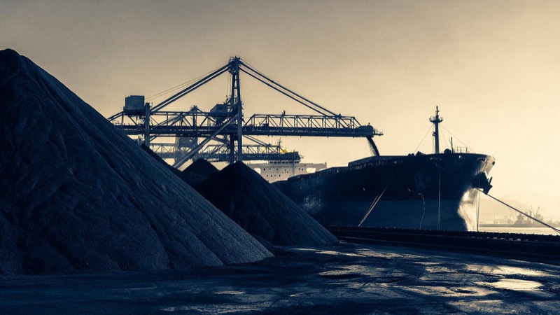 A bulk carrier loads copper concentrate beside towering stockpiles at a Chilean port at dawn, conveyor gantries stretching over the dark hull