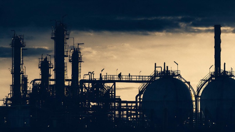 At dusk a Gulf Coast refinery glows amber, its towers, pipes and spherical storage tanks black against a darkening sky, a lone worker's silhouette on a high catwalk