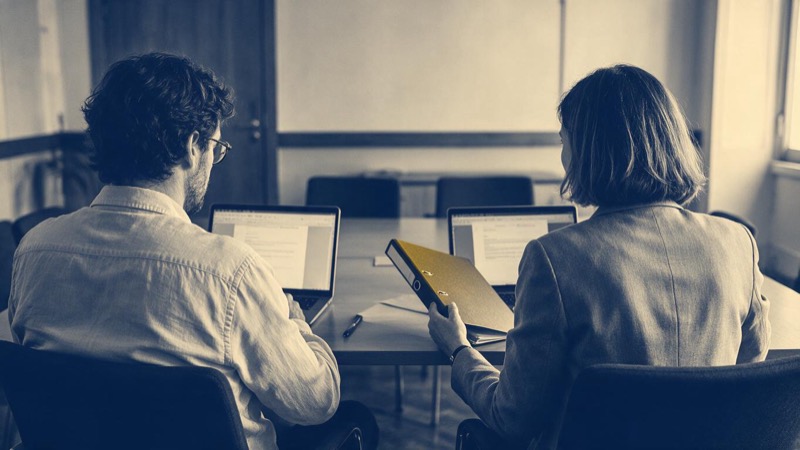 Two auditors seen from behind sit across a meeting-room table in Lisbon, laptops open and one holding a closed binder