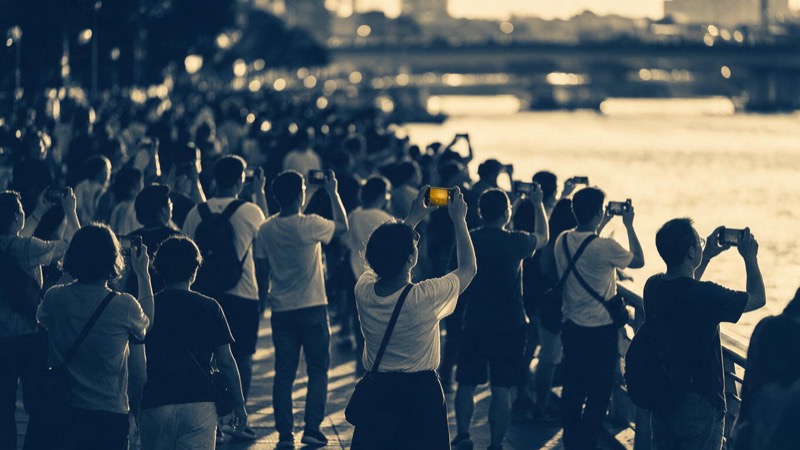 A busy riverside promenade in Porto at sunset with dozens of people holding up phones toward the sky, long gold light on the water