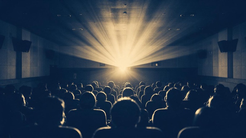 A half-filled cinema auditorium seen from the back row in Seoul, dark silhouettes of the audience's heads below a long beam of dusty projector light