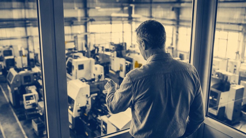 A factory energy manager seen from behind in a glass-walled office overlooking a production hall, one hand pointing down at the machines