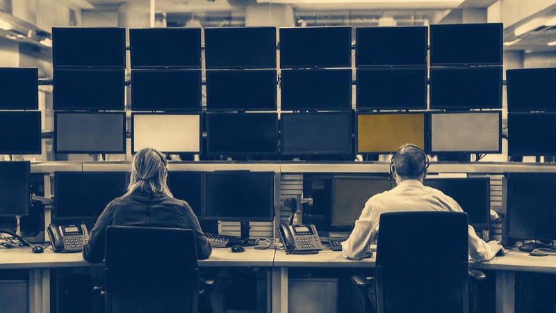 A control room at a stock exchange in London, a wall of dim monitors above desks where two operators sit, seen from behind