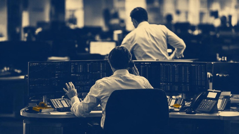 Wide view of a broker's dealing room in New York before the open, long rows of desks with multiple monitors and only a few traders in so far