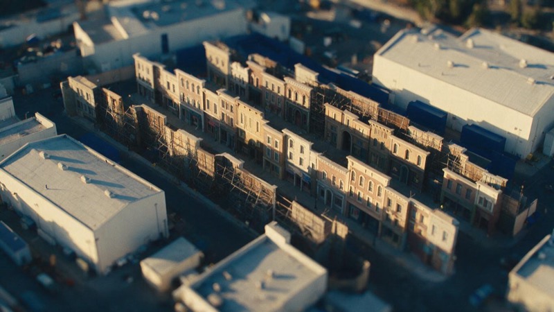 Aerial view of a backlot of false-front street sets and white sound stages at golden hour, scaffolding visible behind the painted facades