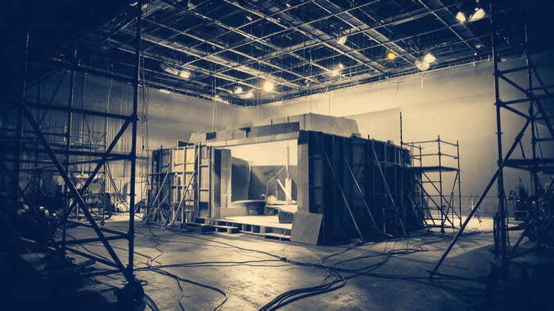 Interior of a vast sound stage with an unfinished set, scaffolding, cables and a lighting grid overhead, work lights on