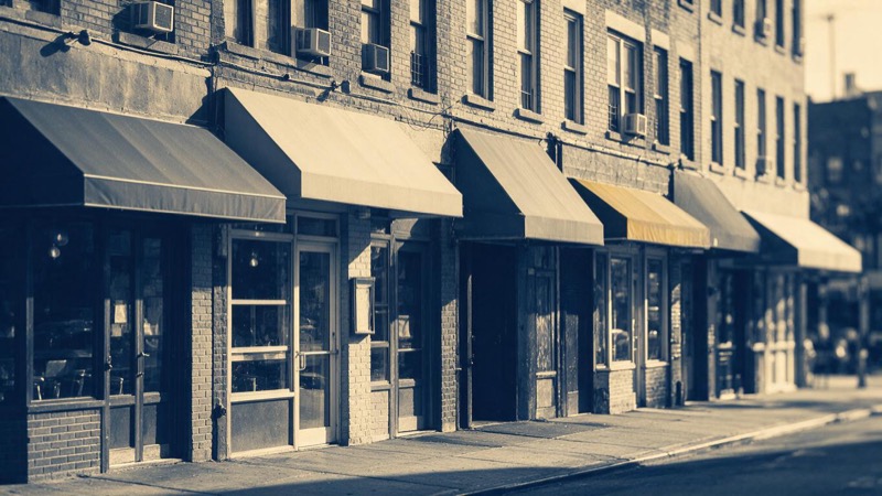 Row of restaurant and café frontages with plain awnings and brick facades on a Brooklyn commercial street in morning light, a few units dark