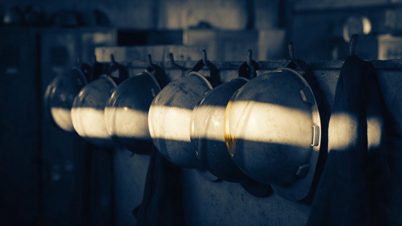 A row of worn hard hats hanging on pegs in a plant locker room, each scuffed and dusty, a single bar of light falling across them