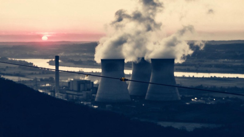 Cooling towers of a nuclear power station releasing steam against a pink sunrise over a flat river valley, a single transmission line crossing the foreground
