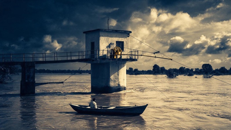 A small concrete river-gauging hut with a cable and winch hanging over a swollen brown river in northern India, monsoon light and a lone boat downstream