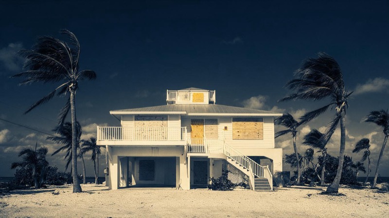 A Florida Keys beach house with plywood boards screwed over every window stands under a darkening sky, palm trees bent by the first gusts of wind