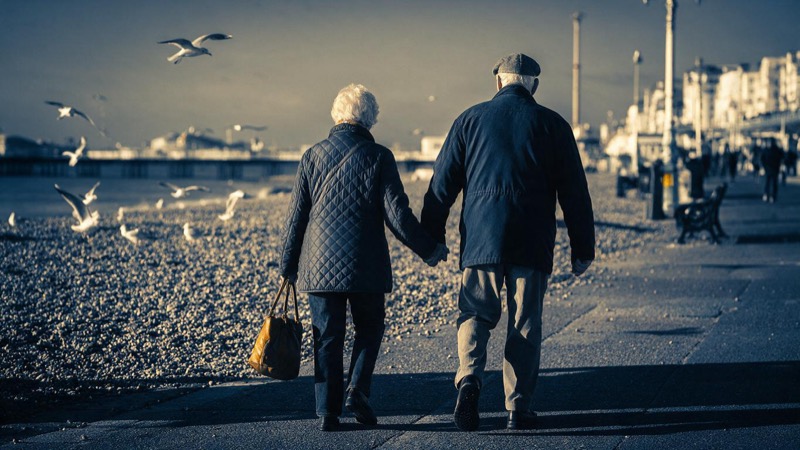 Seen from behind, an elderly couple walk hand in hand along a Brighton seafront promenade on a grey winter morning, gulls over the pebbles