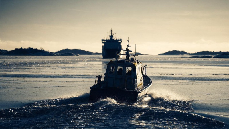 A pilot boat heading out across icy grey water toward the dim silhouette of an approaching ship in winter light, low islands dotted along the Finnish coast