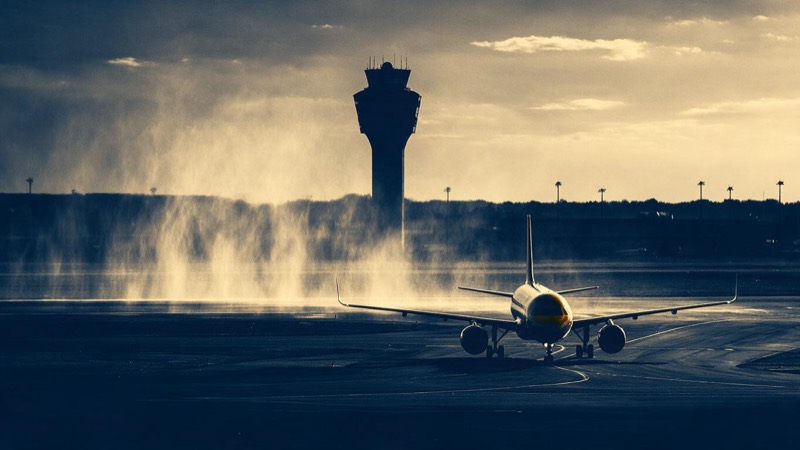An airport control tower silhouetted against a bruised purple sky at dusk, rain curtains approaching across the runway and one aircraft waiting on the taxiway