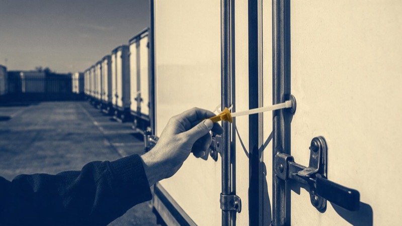 A hand fastening a plastic seal across the rear doors of a white trailer in a lorry yard under flat midday light, rows of identical trailers stretching away to a distant fence