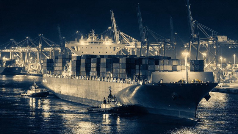 Two small tugboats nudging a huge container ship toward its berth at night, seen from the quay with floodlit cranes towering against a black sky