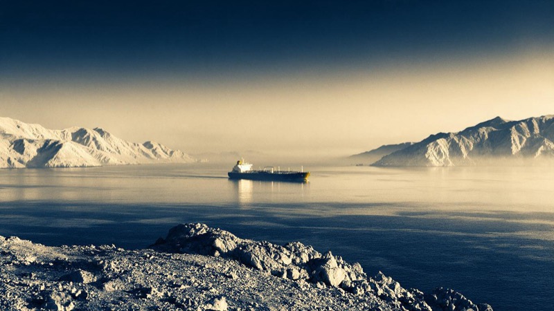 From a rocky headland at sunrise, a single laden tanker crosses a narrow strait flanked by pale desert mountains, haze lying over flat water