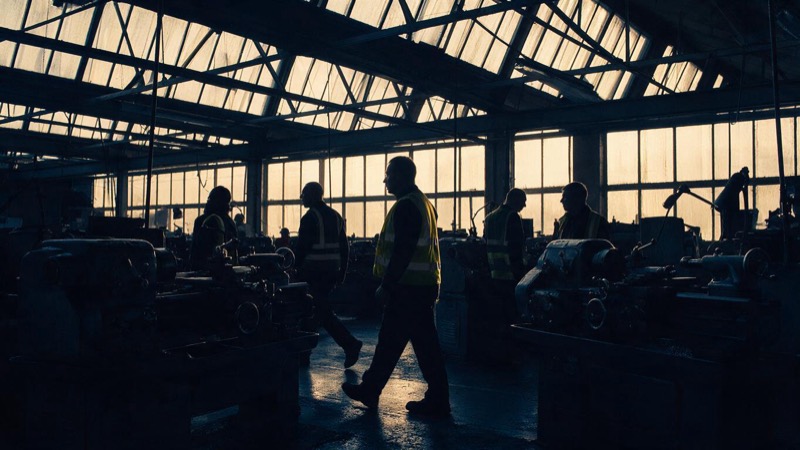 A Lancashire factory floor at shift change, hi-vis silhouettes walking between idle lathes in late-afternoon light falling through sawtooth roof windows