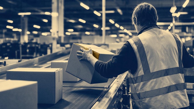 Inside a retail distribution centre in Poland, a conveyor carries plain cartons past a worker seen from behind reaching to lift one off, under harsh strip lights