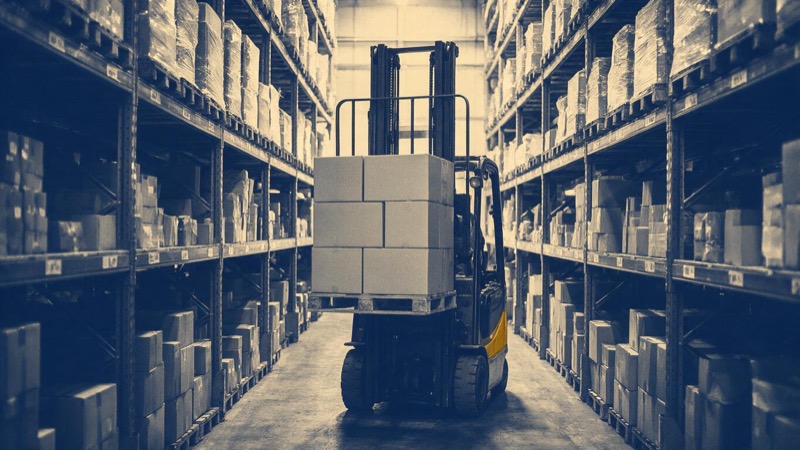 A forklift raising a pallet of plain boxed goods between towering warehouse racks, shot from the far end of the narrow aisle in dim light