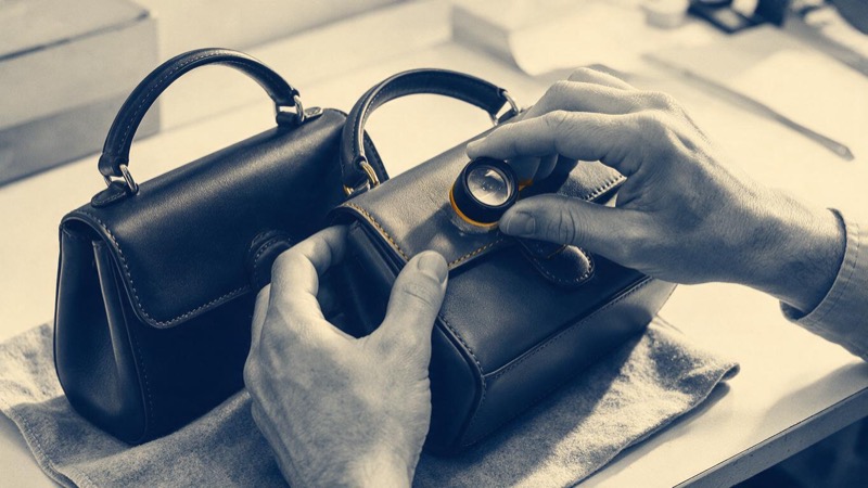 A brand-protection inspector's hands examining the stitching of a leather handbag on a felt cloth with a magnifying loupe, an identical bag beside it, in bright light