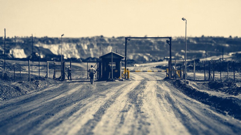 The entrance gate of a copper mine in Zambia's Copperbelt, a security hut and boom gate at the end of a red dirt haul road leading toward the distant pit, midday