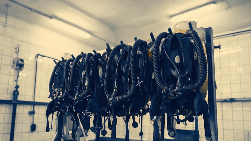 A row of breathing apparatus units hung on a rack in a clean white-tiled mine-rescue training bay, soft overhead light, shot from a low angle
