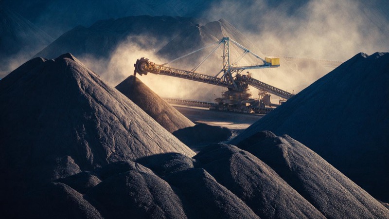 Huge stockpiles of red iron ore at a port stockyard in the Pilbara at dawn, a stacker-reclaimer on rails between the heaps and dust catching the first light