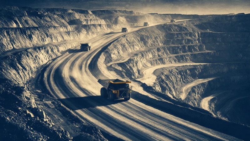 Haul trucks climbing a switchback ramp out of a deep gold pit in Nevada at golden hour, seen from the rim with long shadows across the terraced walls