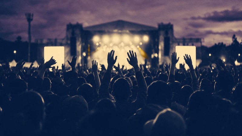 Wide view from the back of a festival crowd in Bogota at dusk, silhouettes with raised hands facing a distant lit stage under a purple sky