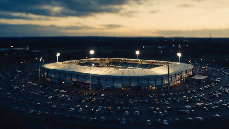 Aerial of a large outdoor arena at dusk with the floodlights on and the car park filling around it